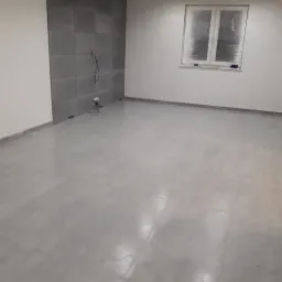 Szare płytki podłogowe i fragment ściany z szarymi panelami w pustym, odświeżonym pokoju z zabezpieczonym okienkiem.