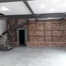 Surowe wnętrze z odsłoniętą cegłą i drewnianymi belkami, spiralne schody z metalową konstrukcją, szara posadzka, widoczne elementy instalacji pod sufitem.