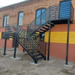 Zewnętrzne schody metalowe z poziomym wypełnieniem balustrady, prowadzące do drzwi na piętrze budynku z cegły i pomalowanego na żółto-pomarańczowo. Schody osadzone na betonowych fundamentach.