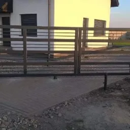 Nowoczesna, pozioma brama wjazdowa z metalowymi przęsłami na posesji z kostki brukowej, widoczny fragment ogrodzenia z siatki i dom w tle.