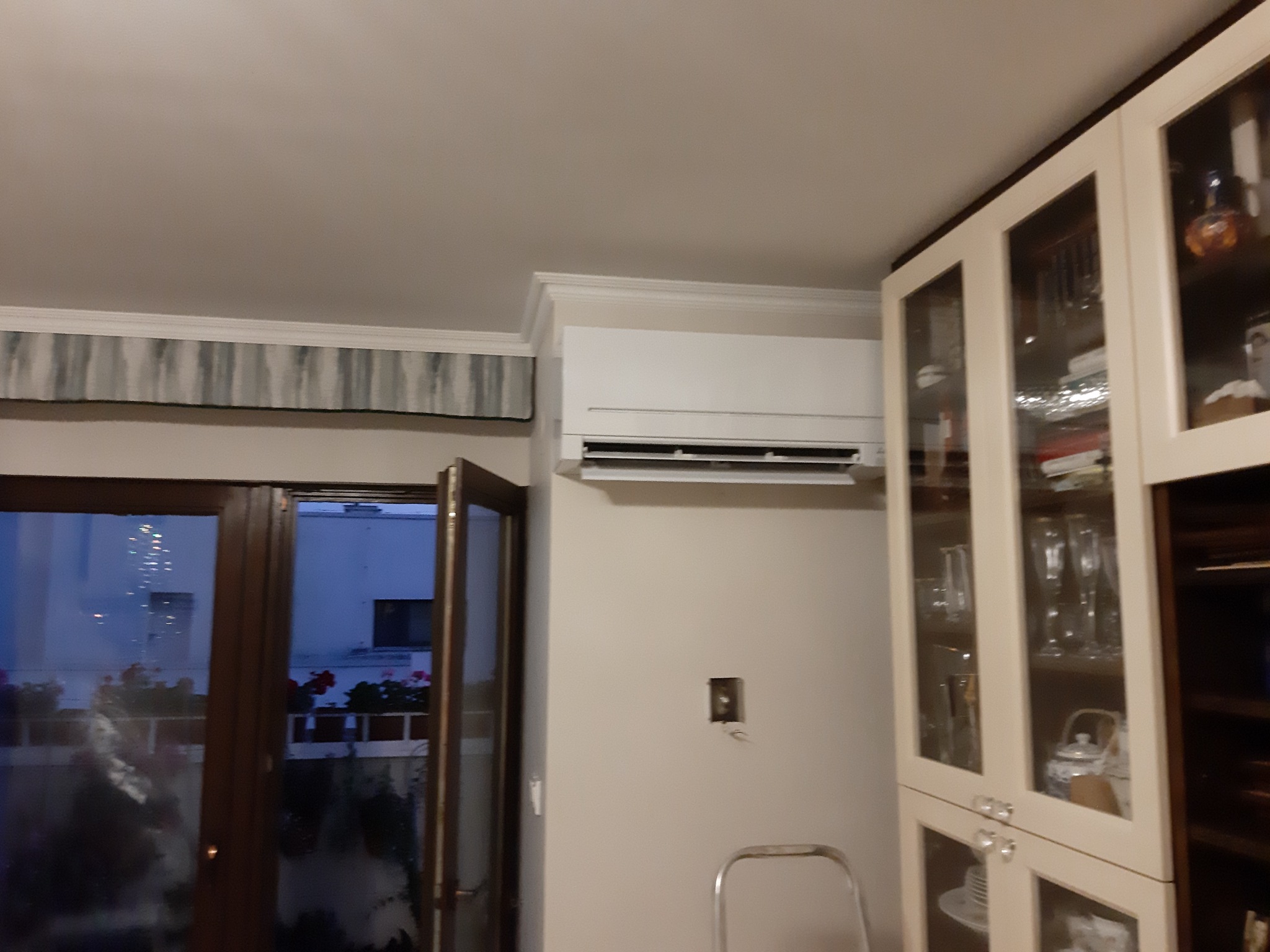 Biała klimatyzacja ścienna w jasnym wnętrzu, widoczna otwarta balkonowa zieleń, obok przeszklona witryna z zastawą.