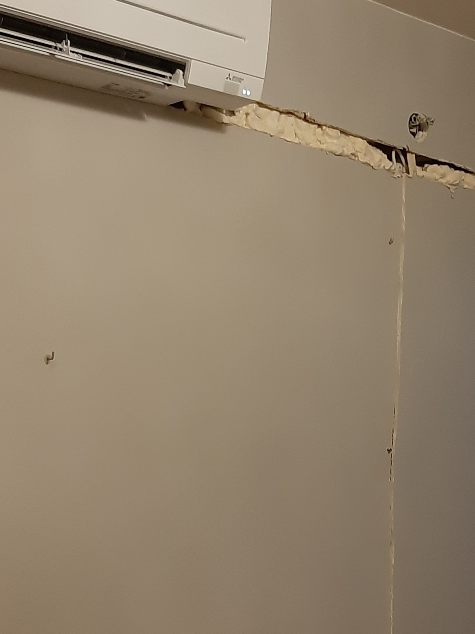 Biały klimatyzator zamontowany na ścianie, widoczne szczeliny uszczelnione pianką montażową i wystający kabel elektryczny.