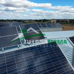 Instalacja paneli słonecznych na dachu budynku z widocznym logo firmy 'Zielona Firma' w odbiciu na panelach, na tle wiejskiego krajobrazu i błękitnego nieba z chmurami.