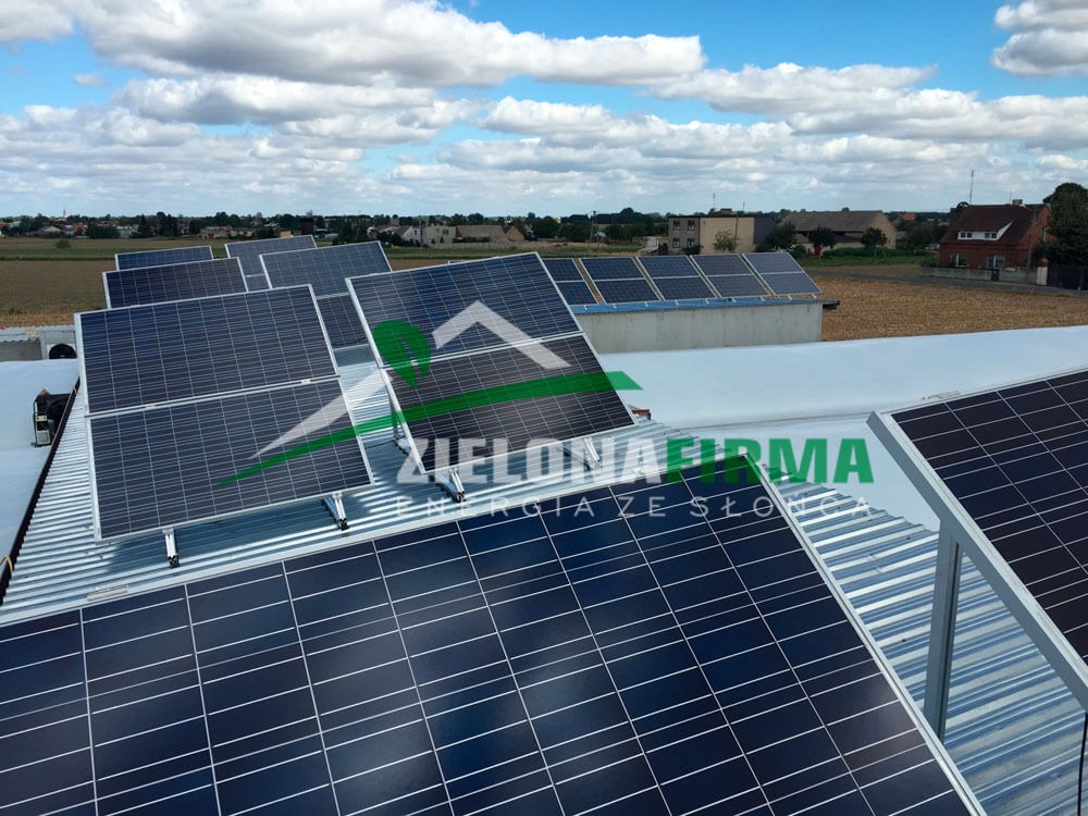 Instalacja paneli słonecznych na dachu budynku z widocznym logo firmy 'Zielona Firma' w odbiciu na panelach, na tle wiejskiego krajobrazu i błękitnego nieba z chmurami.