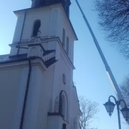 Kościół z wieżą i krzyżem, podnośnik koszowy przy dachu, widok z dołu w słoneczny dzień.