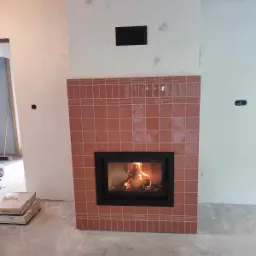 Wnętrze w trakcie wykończenia z obudowanym w różowe płytki wkładem kominkowym z palącym się ogniem.