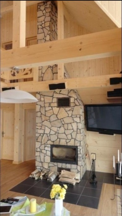 Rustykalny salon z drewnianymi ścianami i kamiennym obłożeniem kominka, nad którym widać fragment kamiennego komina. Na czarnej posadzce leżą przygotowane do palenia kawałki drewna, a obok stoją...