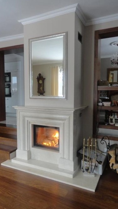 Elegancki, biały portal z płonącym paleniskiem, nad nim lustro w srebrnej ramie, odbijające zabytkowy zegar i zasłony. Podłoga z ciemnego drewna, obok akcesoria kominkowe.