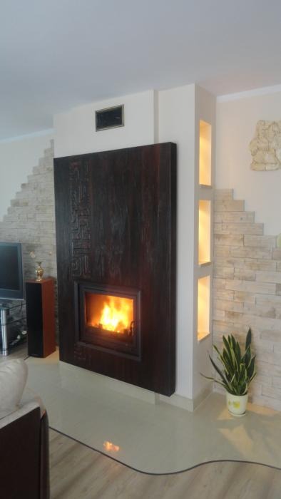Nowoczesny, ciemnobrązowy wkład kominkowy z płomieniami, obudowany płytą z delikatnymi, orientalnymi wzorami, wkomponowany w jasne wnętrze z dekoracyjną ścianą z kamienia i wnękami oświetleniowymi.