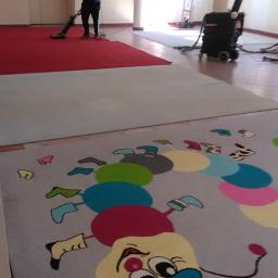 Jg pranie - Pracownik w czarnym uniformie odkurza czerwony dywan w przestronnym pomieszczeniu; w tle widoczny sprzęt do czyszczenia, a na pierwszym planie dywan z kolorowym wzorem kreskówkowym.