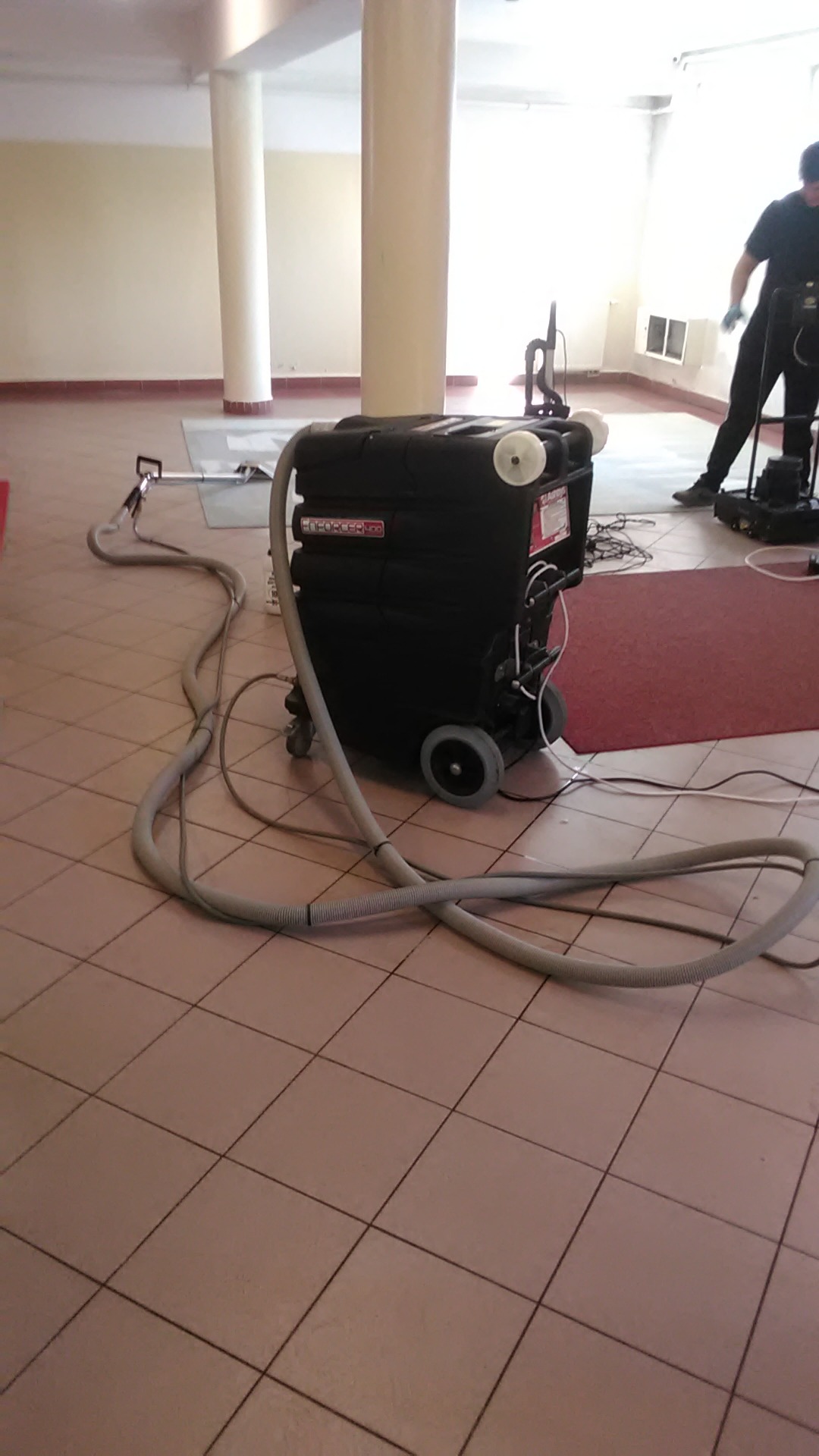 Profesjonalny sprzęt do czyszczenia dywanów marki Enforcer 500 na kafelkowej podłodze w Strzelinie, w tle osoba obsługująca mniejszą maszynę czyszczącą.