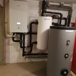 Zainstalowana pompa ciepła Panasonic z dwoma zbiornikami na wodę, widoczne rury i zawory w kotłowni wyłożonej kafelkami.