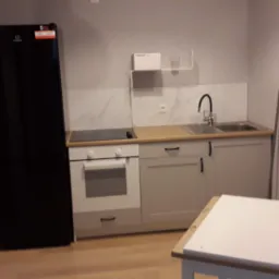 Wyposażona kuchnia z czarną lodówką, jasnymi szafkami i białym stołem, widok na płytki imitujące marmur nad blatem.