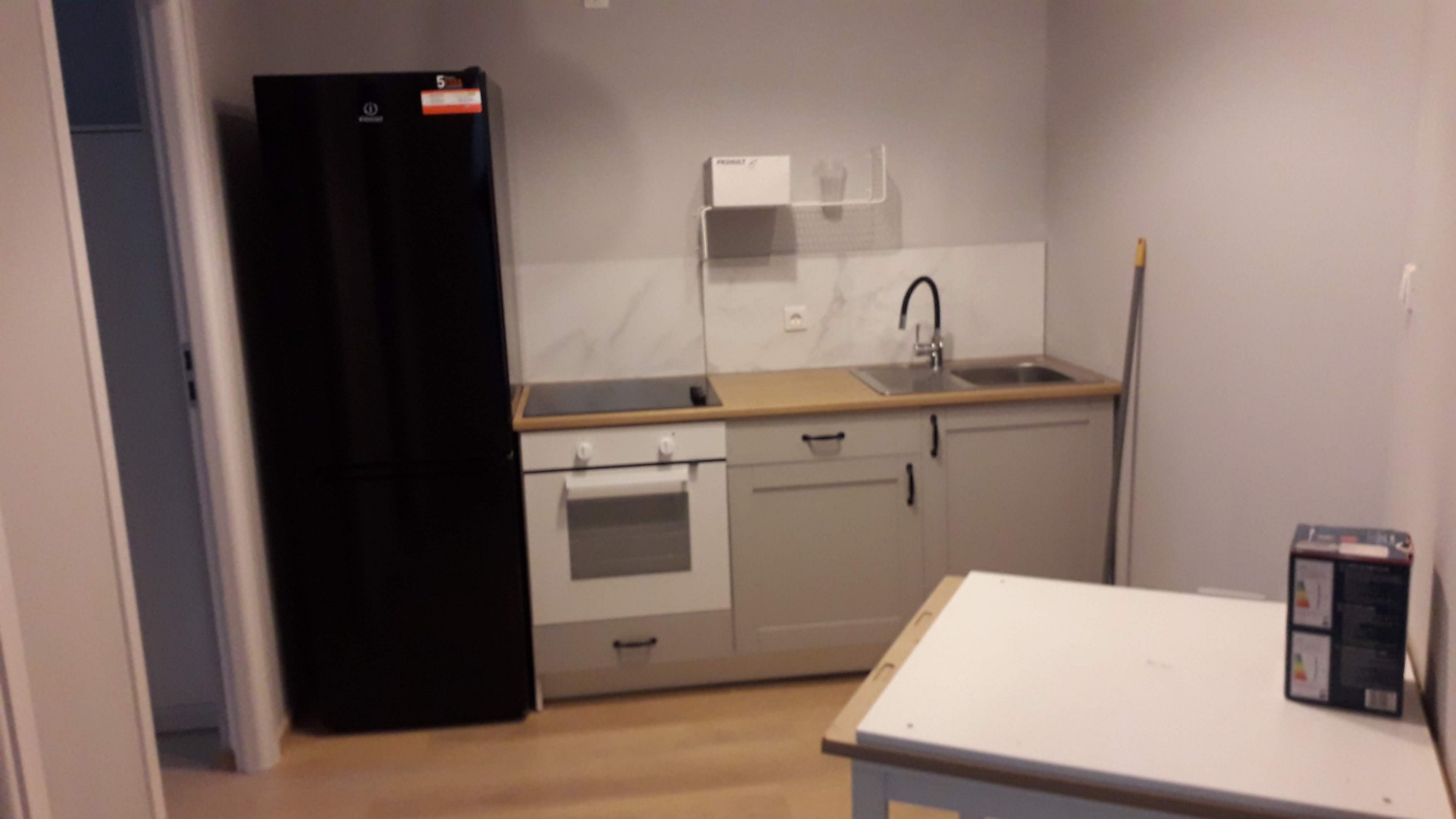 Wyposażona kuchnia z czarną lodówką, jasnymi szafkami i białym stołem, widok na płytki imitujące marmur nad blatem.