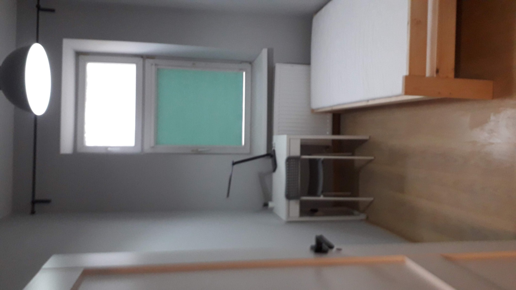 Pokój po remoncie: Świeżo pomalowane ściany w odcieniach szarości, okno z zieloną roletą, nowoczesna lampa wisząca i meble do zabudowy. Widoczne ślady po szpachlowaniu na ścianie w tle.