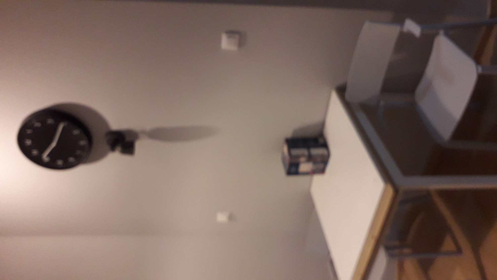 Czarny zegar ścienny z białymi wskazówkami, zamontowany na szarej ścianie w pobliżu białego sufitu z widocznym czujnikiem dymu i częściowo widocznym narożnikiem ściany oraz mebli.