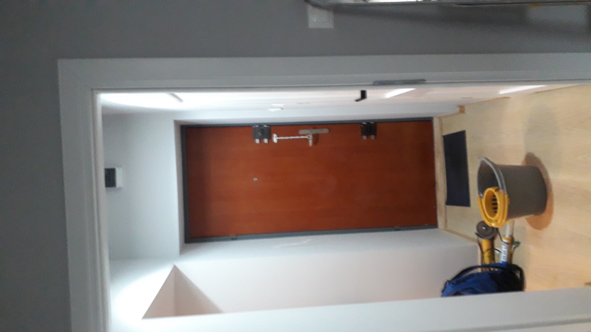 Widok przez framugę drzwi na brązowe drzwi z zabezpieczeniem łańcuchowym, obok wiadro z mopem i narzędzia na podłodze