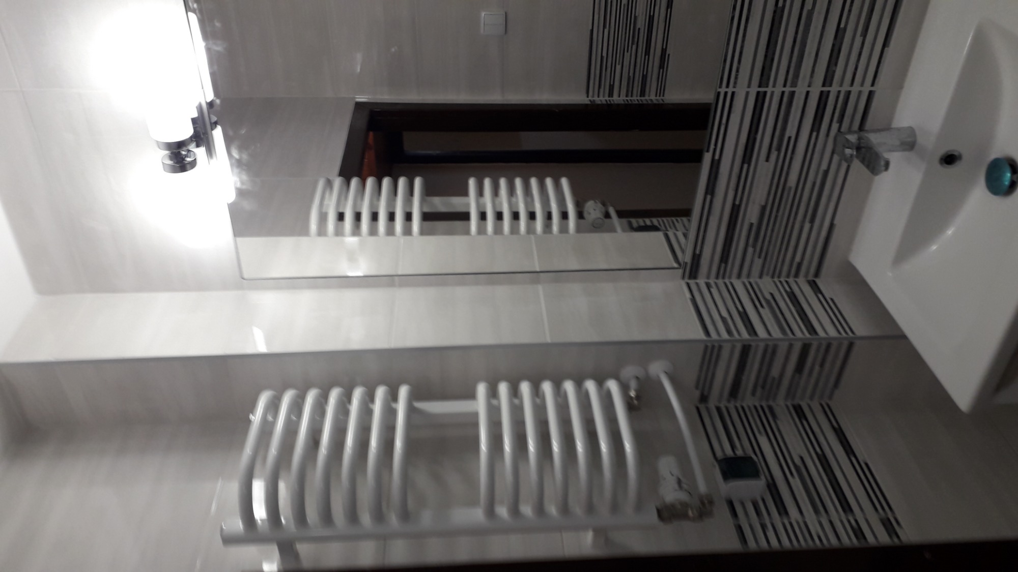 Biała łazienka z grzejnikiem drabinkowym i umywalką, wykończona płytkami w odcieniach szarości z motywem pasków, odbicie w lustrze ukazuje fragment grzejnika i sufitu.