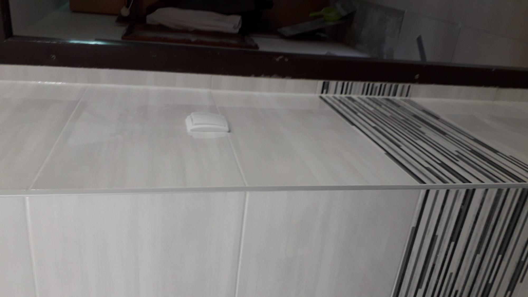 Wykończona ściana i blat z jasnych płytek ceramicznych z ozdobnym pasem czarno-białych pasków, widoczny odpływ liniowy i fragment lustra z brązową ramą.