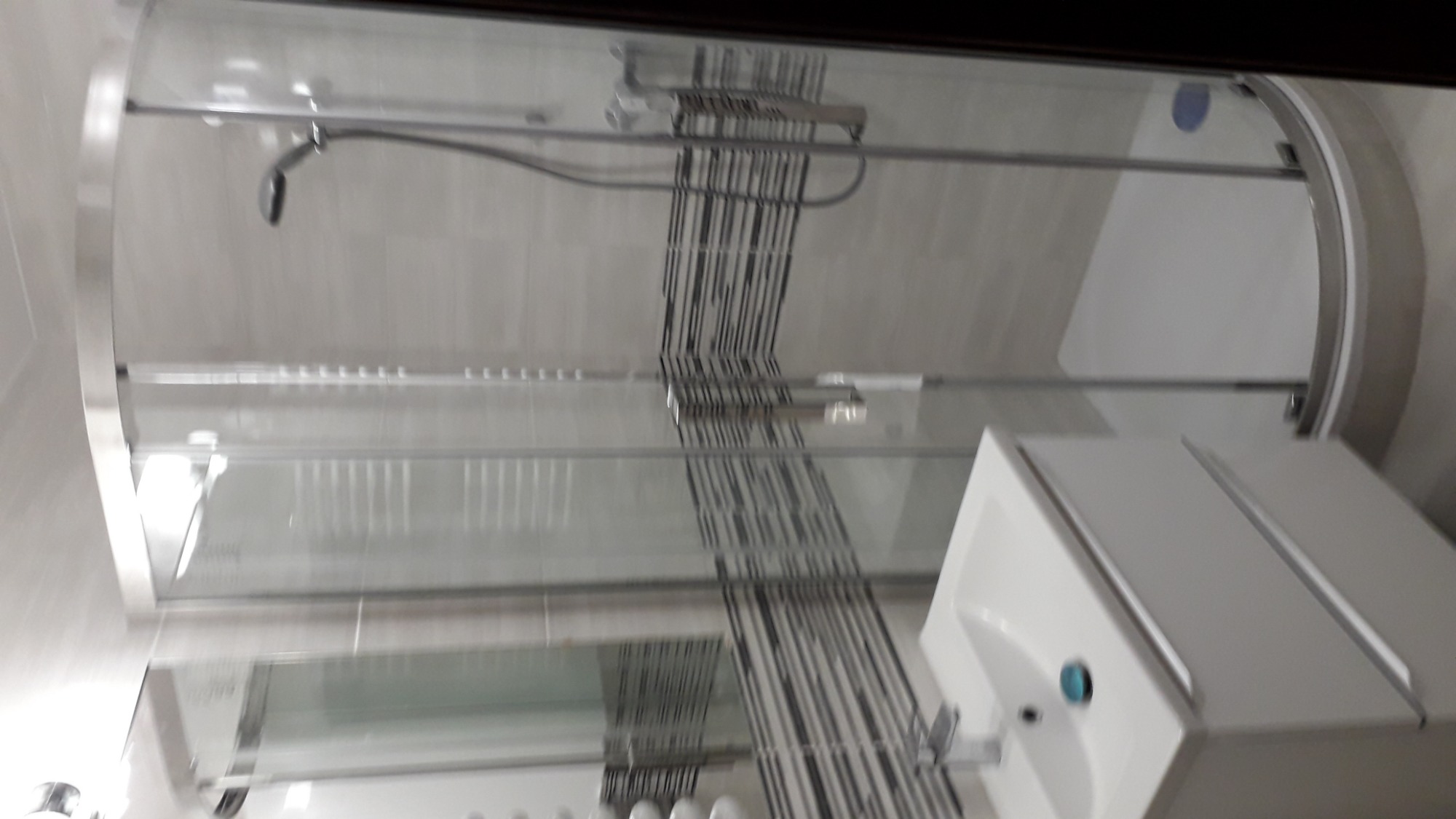 Nowoczesna kabina prysznicowa z chromowanymi elementami, widoczna umywalka z szafką w minimalistycznym stylu, szklane drzwi z dekoracyjnymi pionowymi paskami.