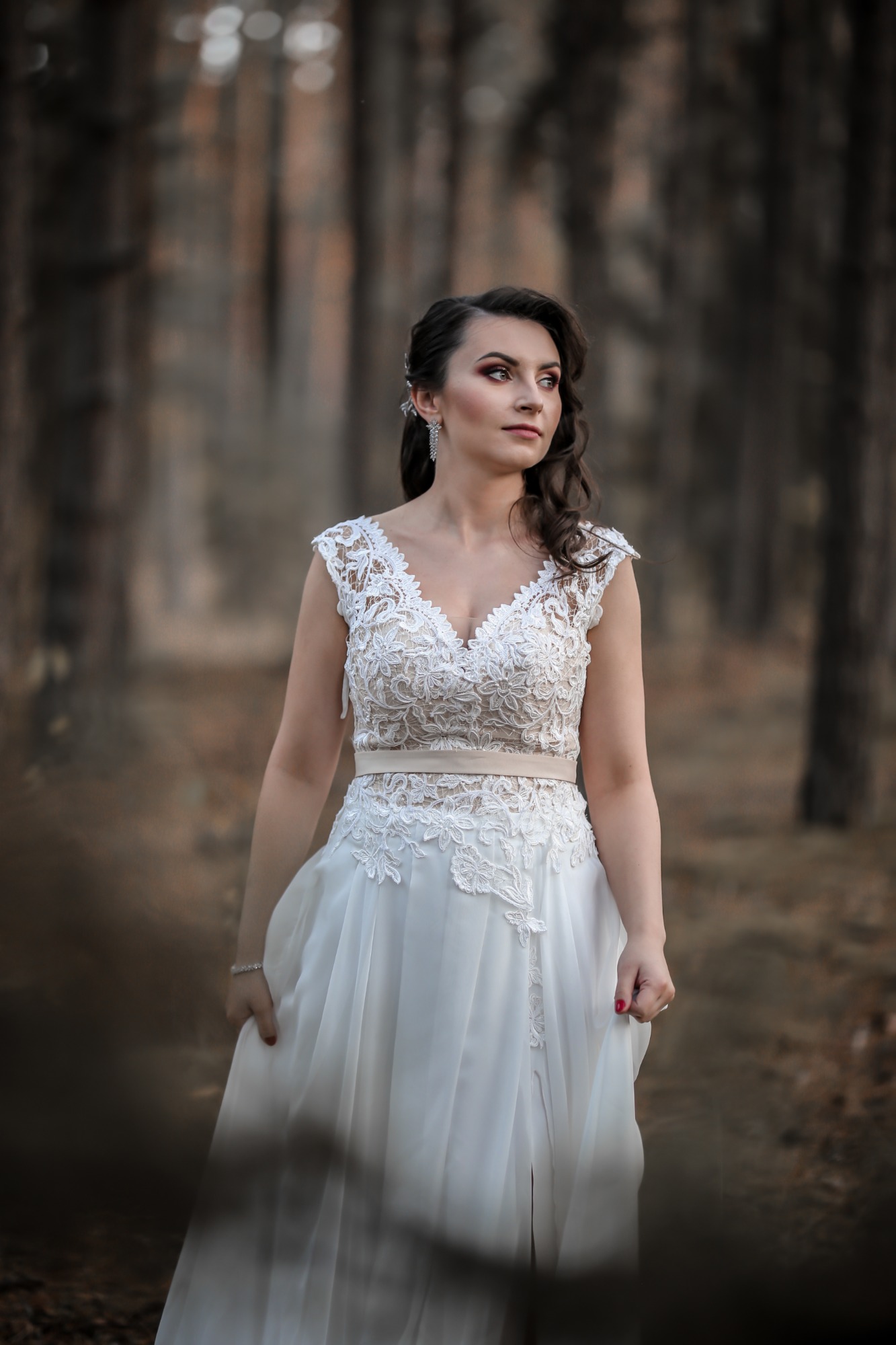 Kobieta w białej sukni z koronkową górą, z upiętymi ciemnymi włosami i delikatnym makijażem, pozuje w lesie, trzymając brzeg sukni.