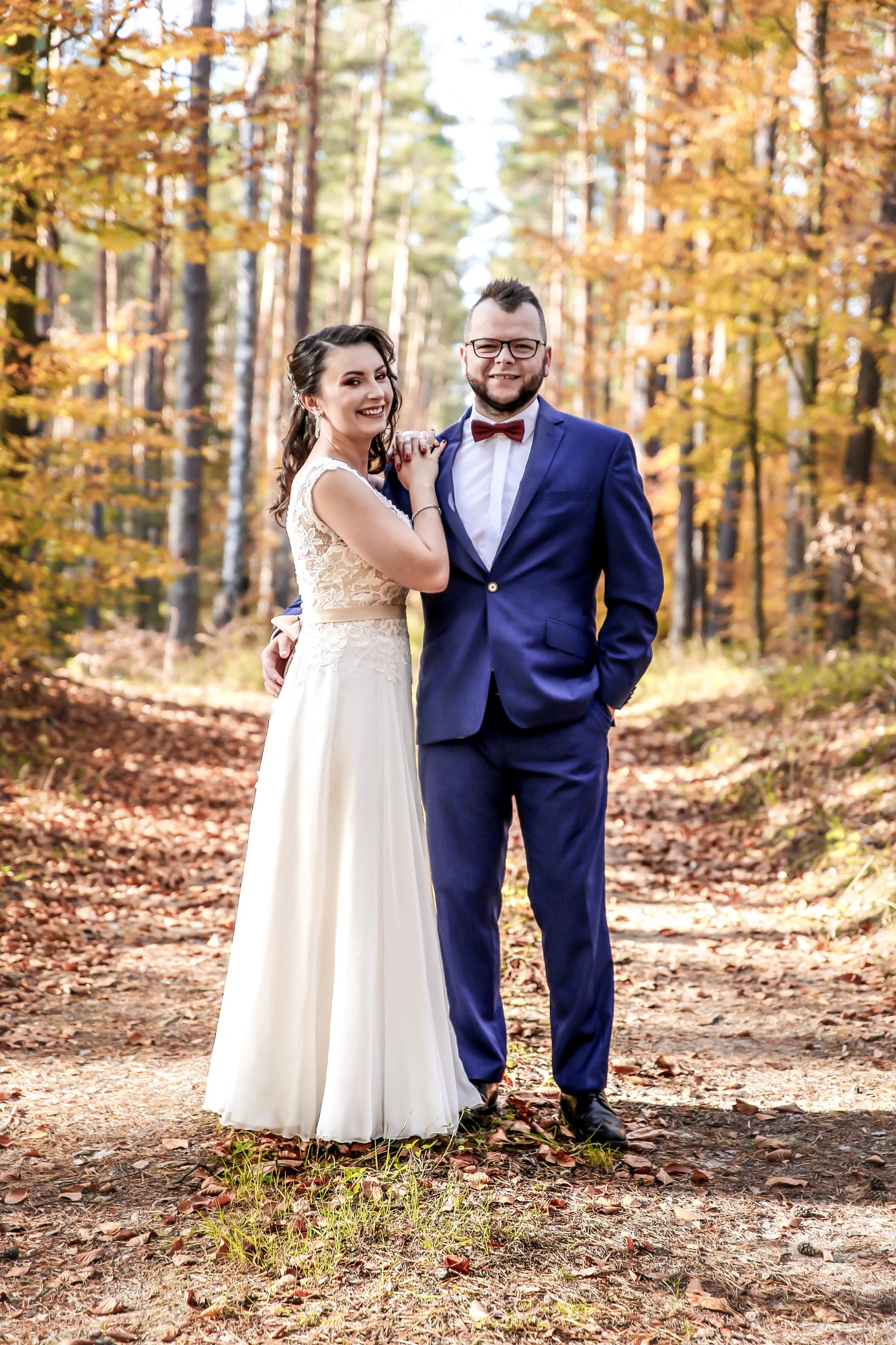 Para w strojach ślubnych pozuje w jesiennym lesie, panna młoda w koronkowej sukni z delikatnym makijażem, pan młody w granatowym garniturze z bordową muchą.