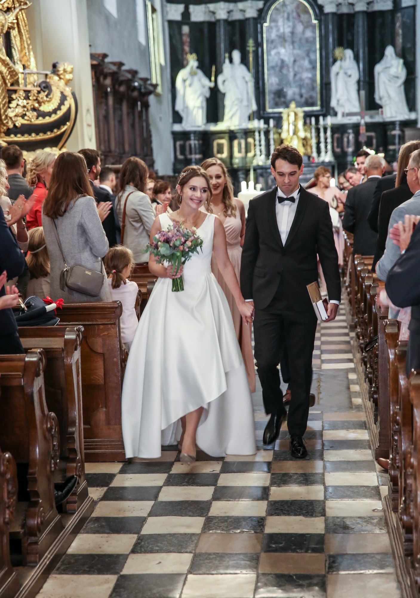 Para młoda, on w czarnym smokingu z muchą, ona w białej sukni z bukietem, idą przez kościół w Niedrzwicy Dużej, oklaskiwani przez gości.