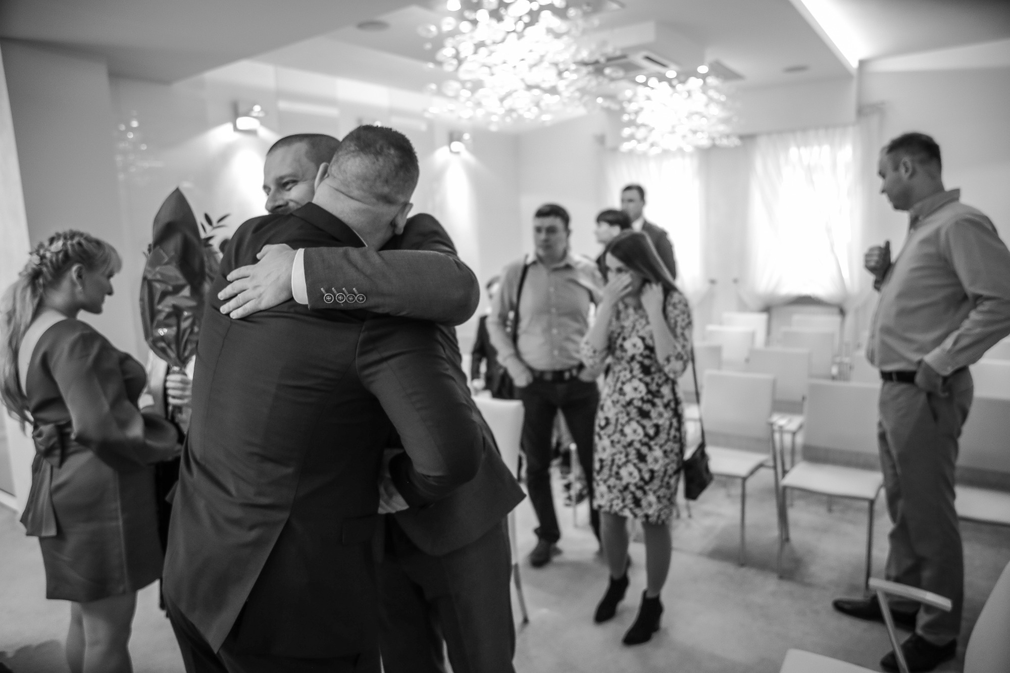 Czarno-białe zdjęcie z uroczystości, mężczyźni w garniturach obejmują się, w tle widoczni goście i dekoracyjne oświetlenie sufitowe.