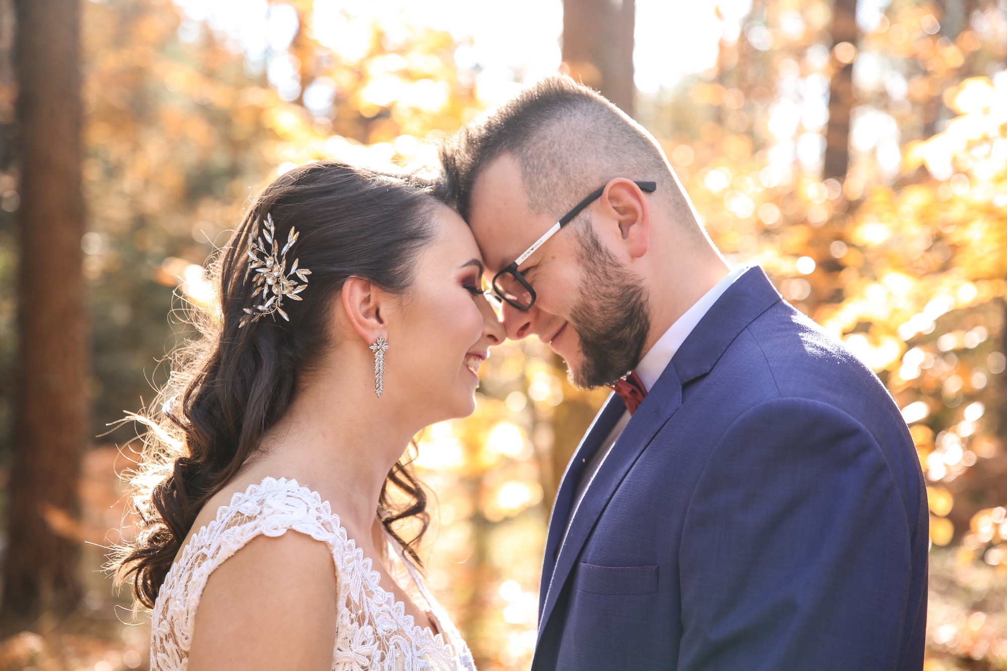 Uśmiechnięta para, panna młoda w koronkowej sukni i ozdobie we włosach, pan młody w niebieskim garniturze i bordowej muszce, dotykają się czołami na tle jesiennego lasu.