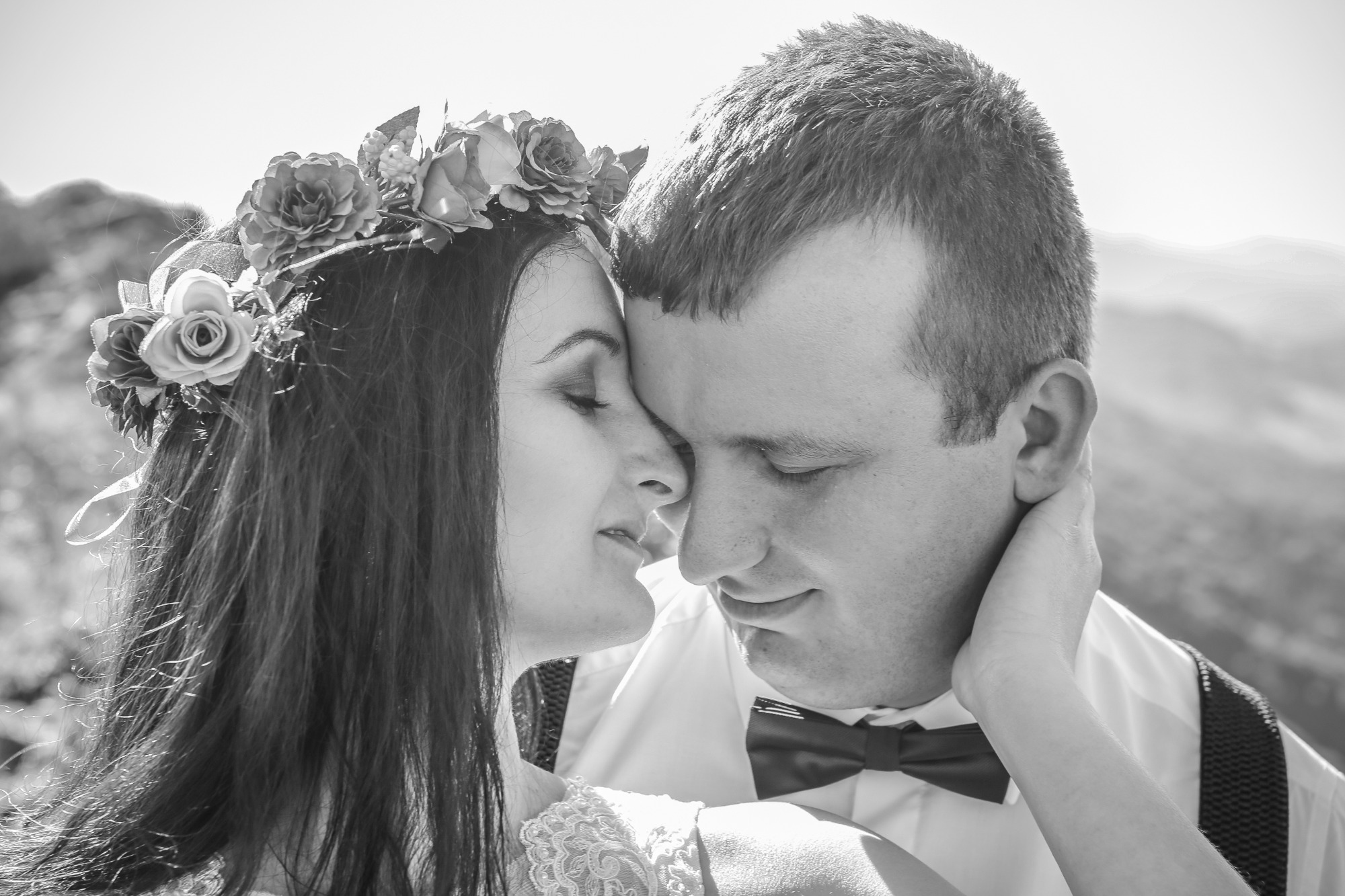 Czarno-białe zbliżenie pary: kobieta z wiankiem z kwiatów na głowie dotyka twarzą do twarzy mężczyzny z muchą, w tle rozmyty krajobraz.