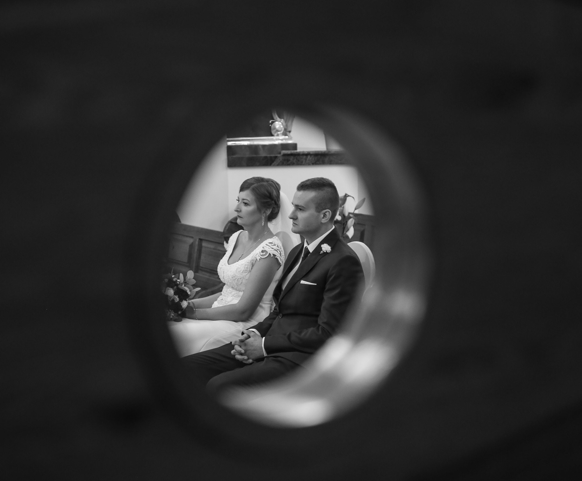 Czarno-białe zdjęcie pary młodej siedzącej w kościele, uchwycone przez okrągły otwór, z widocznymi detalami sukni panny młodej i bukietu.
