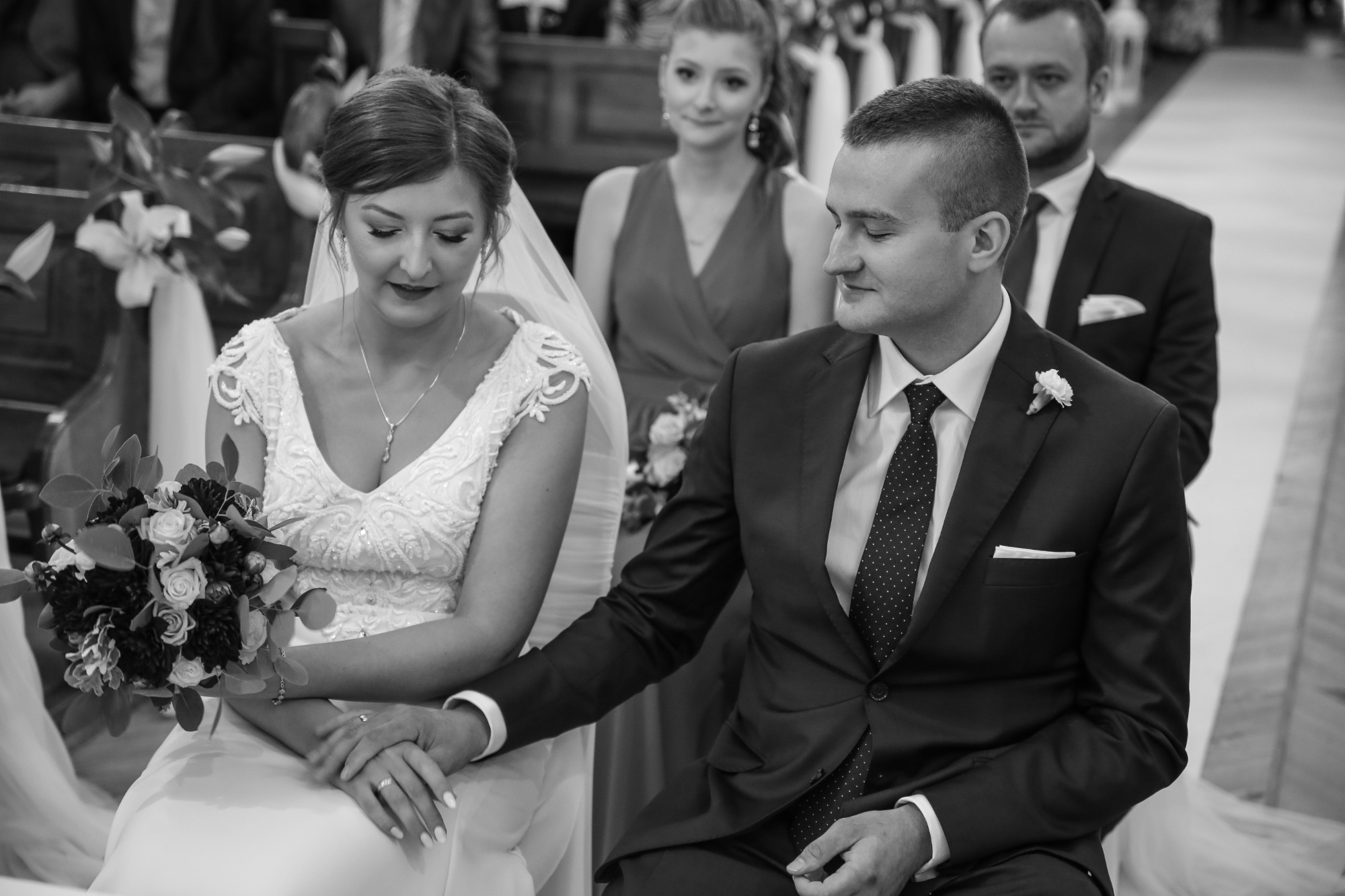 Czarno-białe zdjęcie pary młodej siedzącej obok siebie w kościele, panna młoda trzyma bukiet, a pan młody obejmuje jej dłoń. W tle druhny i goście.