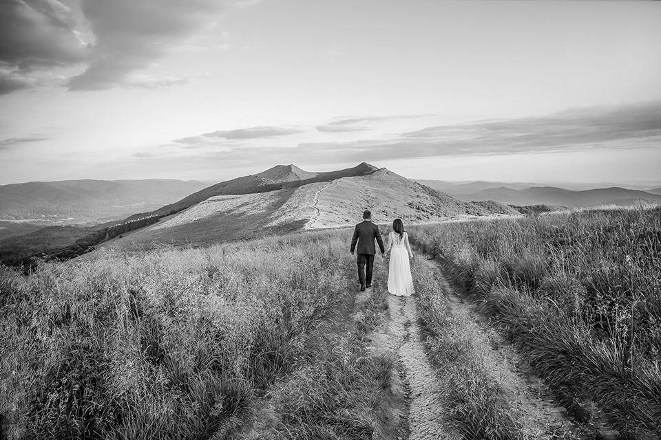 Czarno-białe zdjęcie pary młodej idącej ścieżką na tle górskiego krajobrazu. Mężczyzna w ciemnym garniturze, kobieta w jasnej sukni ślubnej, trzymają się za ręce. W tle widoczny szczyt góry...