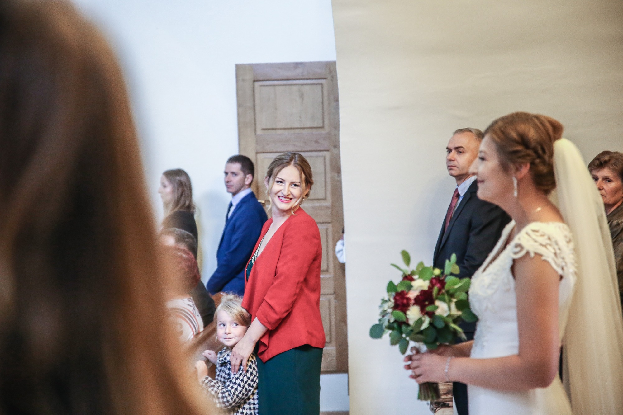 Panna młoda w białej sukni ślubnej trzyma bukiet, obok kobieta w czerwonym żakiecie z dzieckiem, w tle goście weselni.