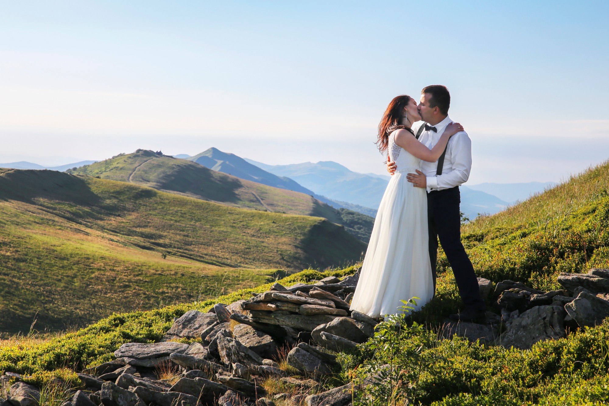Para całująca się na szczycie góry w dniu ślubu, ona w białej sukni, on w koszuli z szelkami i muszką, w tle rozległe pasmo górskie.
