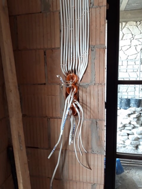 Białe przewody elektryczne prowadzone w ścianie z cegły, widoczne pomarańczowe puszki instalacyjne, etap prac w surowym stanie.