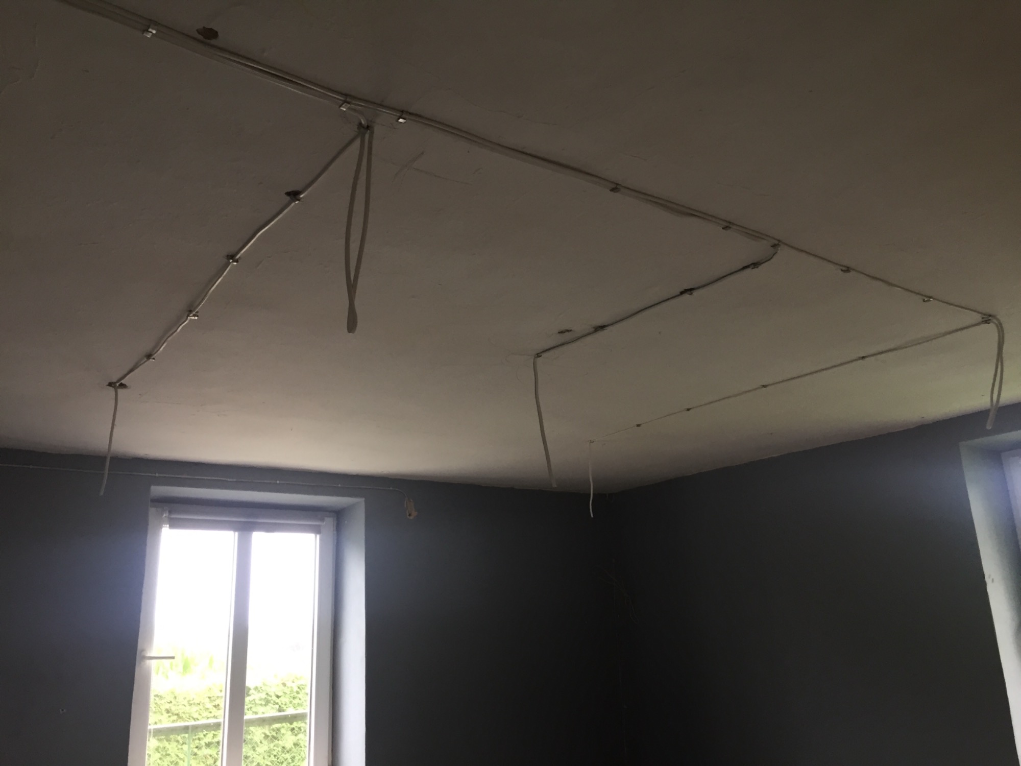 Montaż szyny oświetleniowej na suficie w pokoju z szarymi ścianami, widoczne kable zwisające z sufitu, okno w lewym dolnym rogu.