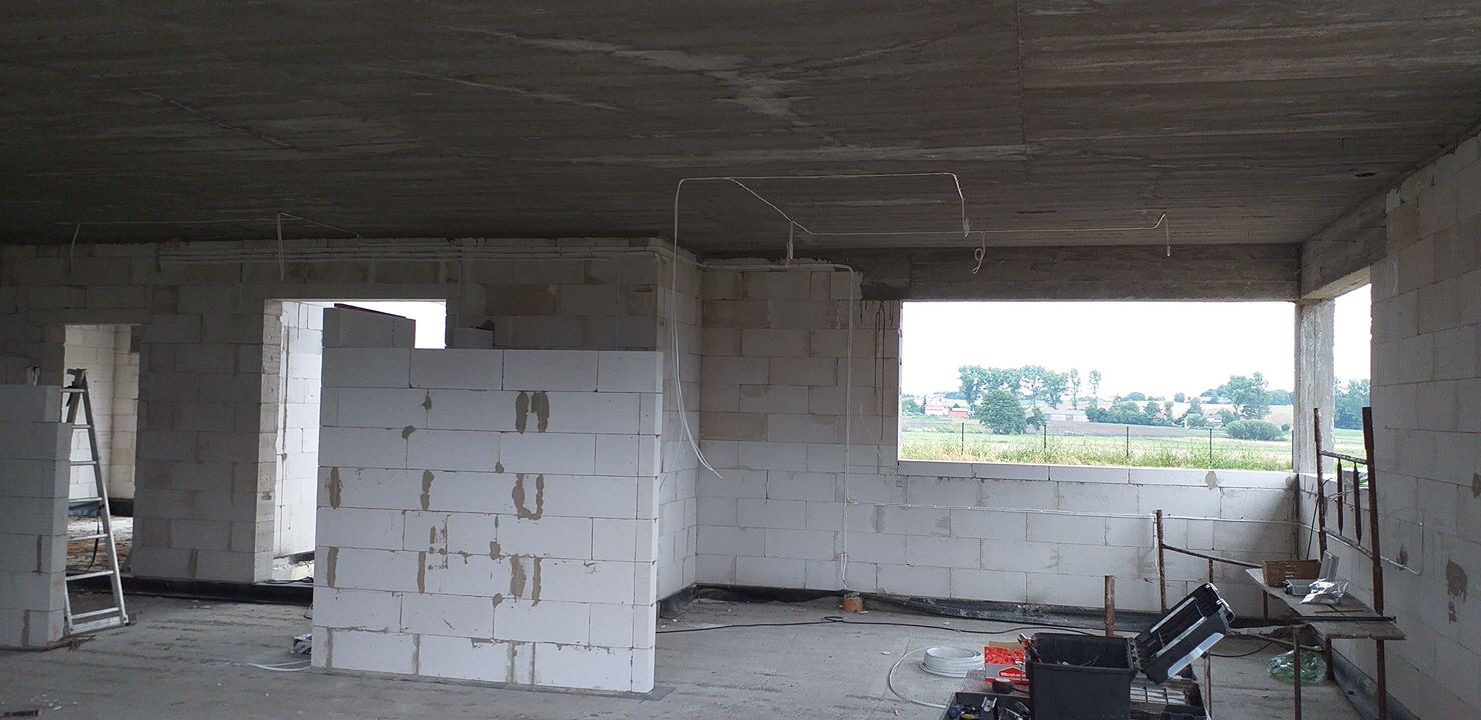 Surowy stan budynku w trakcie prac, widoczne białe ściany z betonu komórkowego, surowy betonowy strop, wystające przewody elektryczne, drabina i narzędzia na podłodze.