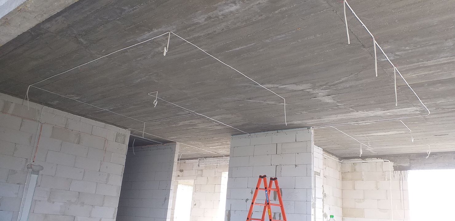 Surowy betonowy strop z widoczną instalacją elektryczną, kable poprowadzone wzdłuż ścian i sufitu, pomarańczowa drabina w tle, wnętrze w stanie deweloperskim.
