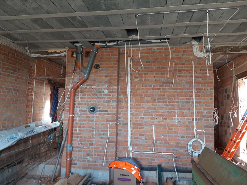 Surowy stan budynku w trakcie prac instalacyjnych: widoczne przewody elektryczne poprowadzone po ceglanej ścianie, rury kanalizacyjne, strop betonowy, oraz elementy rusztowania i narzędzia.