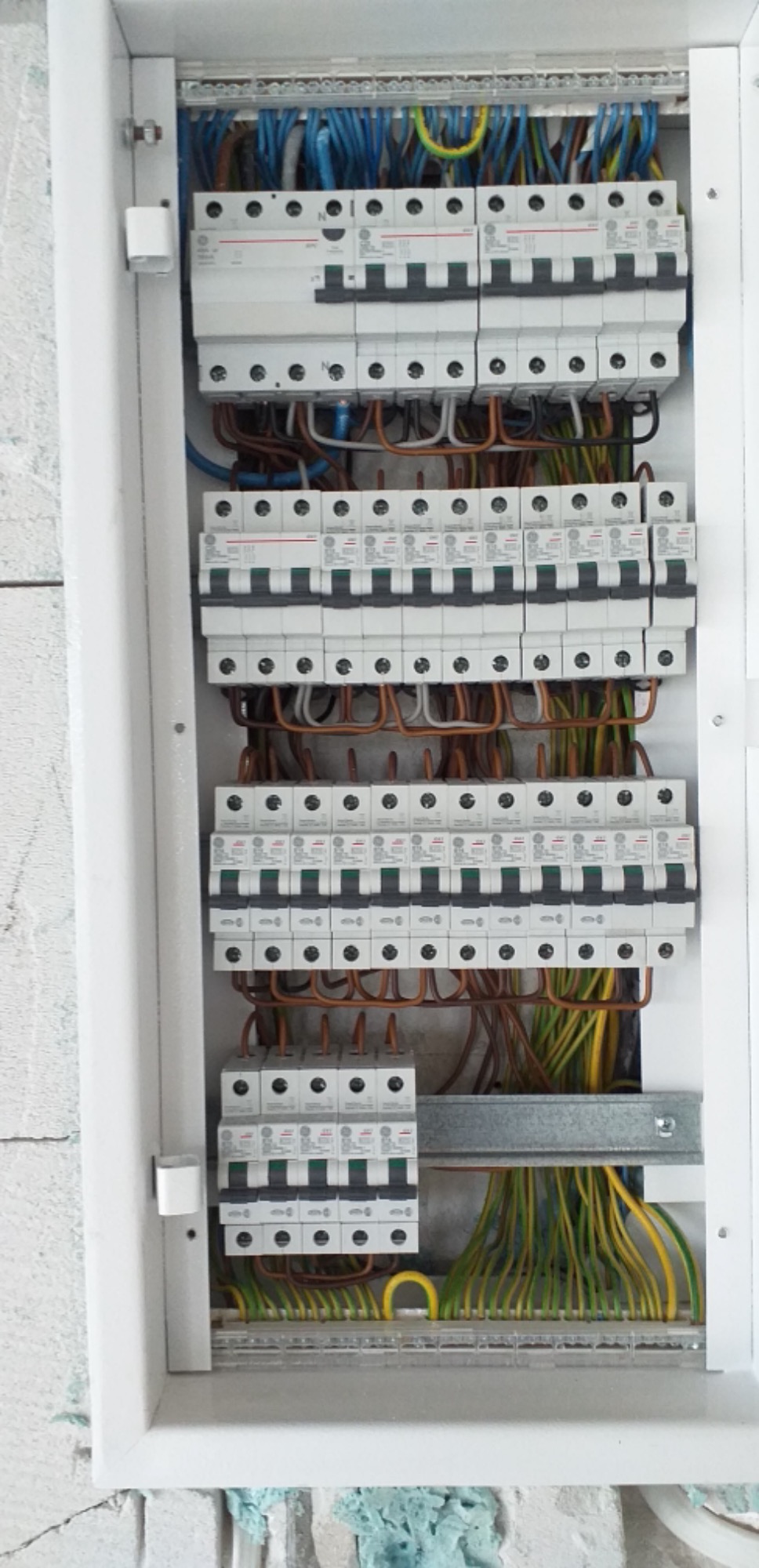 Rozdzielnica elektryczna z zamontowanymi bezpiecznikami i uporządkowanymi kablami w kolorach niebieskim, brązowym i żółto-zielonym, widoczna na tle surowej ściany.