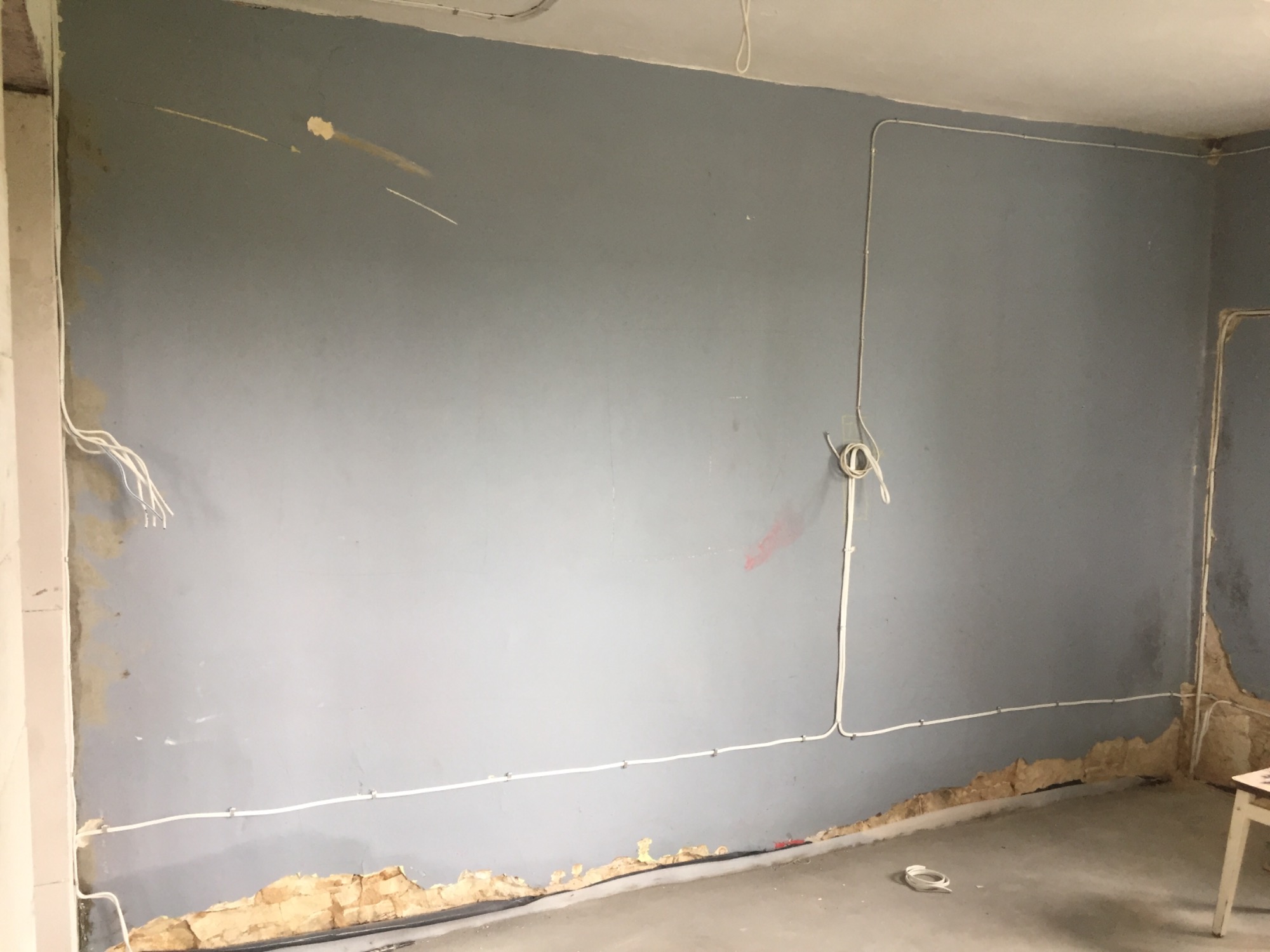 Szare wnętrze w trakcie remontu z widocznymi przewodami elektrycznymi poprowadzonymi po ścianie i pod sufitem, częściowo zakrytymi listwami, oraz odsłoniętym fragmentem ściany przy podłodze.