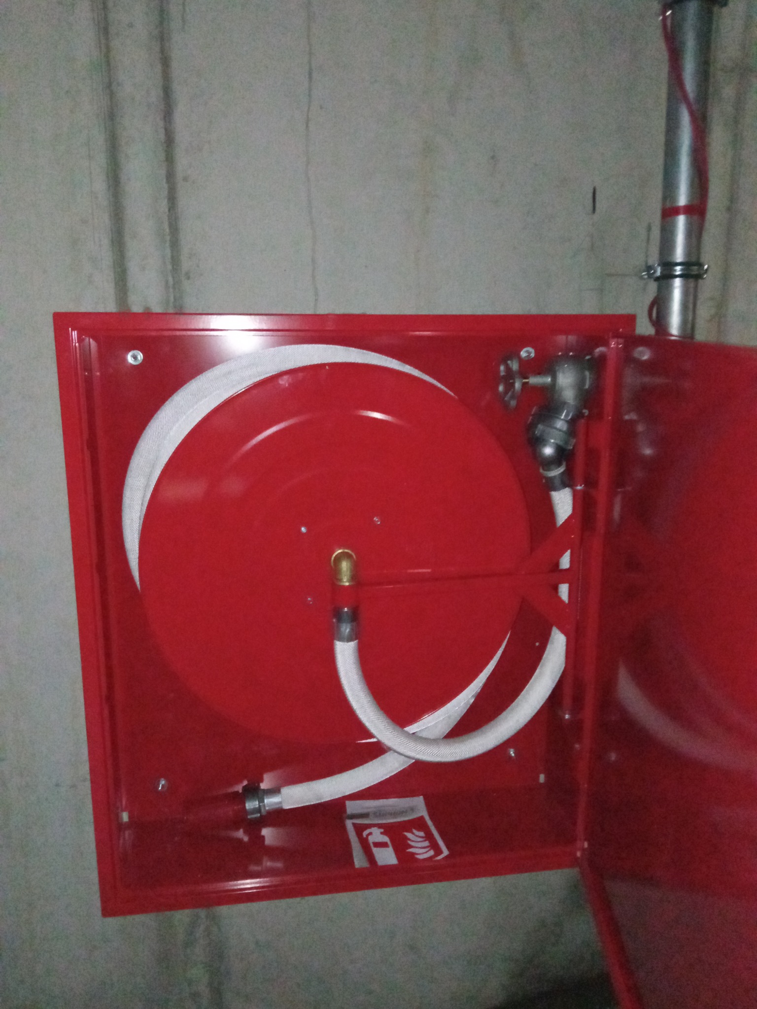 Czerwona szafka hydrantowa z rozwiniętym wężem, zamontowana na szarej, betonowej ścianie wewnątrz budynku.