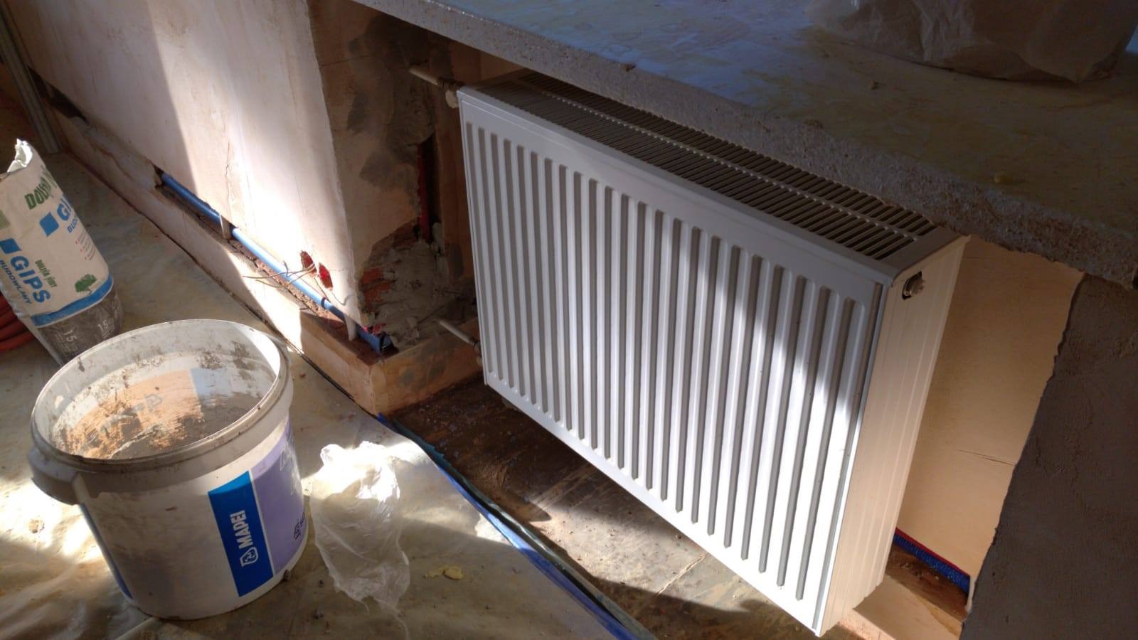 Montaż nowego, białego grzejnika panelowego w trakcie modernizacji instalacji centralnego ogrzewania; widoczne rury instalacyjne w ścianie, wiadro z zaprawą i worek gipsu budowlanego.