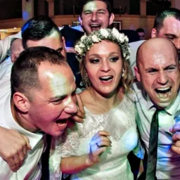 Panna młoda w wianku i otaczający ją mężczyźni śpiewają do mikrofonu podczas wesela, wszyscy z entuzjazmem otwierają usta.