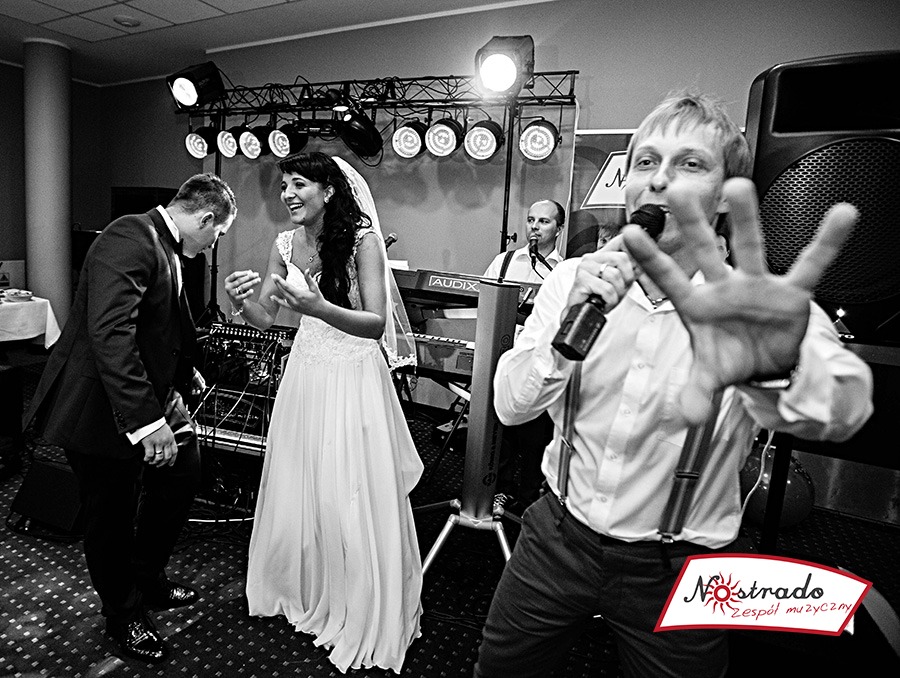 Czarno-białe zdjęcie z wesela: wokalista zespołu muzycznego z mikrofonem na pierwszym planie, za nim para młoda, klawiszowiec i oświetlenie sceniczne.