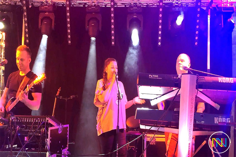 Zespół muzyczny na scenie podczas występu, oświetlony reflektorami punktowymi, z gitarzystą po lewej, wokalistką pośrodku i klawiszowcem po prawej stronie.