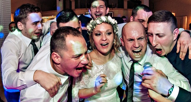 Panna młoda w wianku i otaczający ją mężczyźni śpiewają do mikrofonu podczas wesela, wszyscy z entuzjazmem otwierają usta.