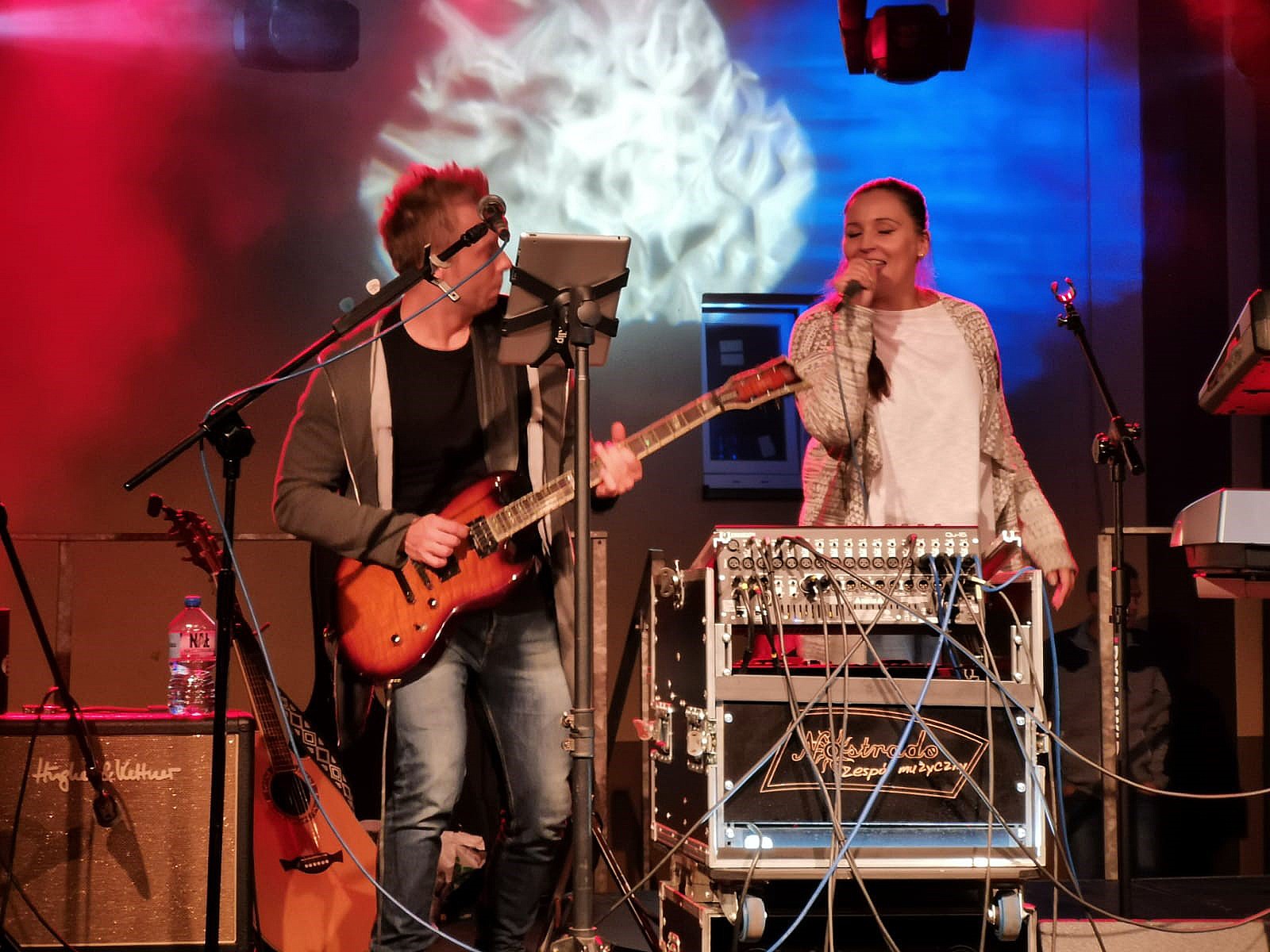 Wokalistka z mikrofonem i gitarzysta z gitarą elektryczną podczas występu na scenie, widoczny mikser i wzmacniacz gitarowy.