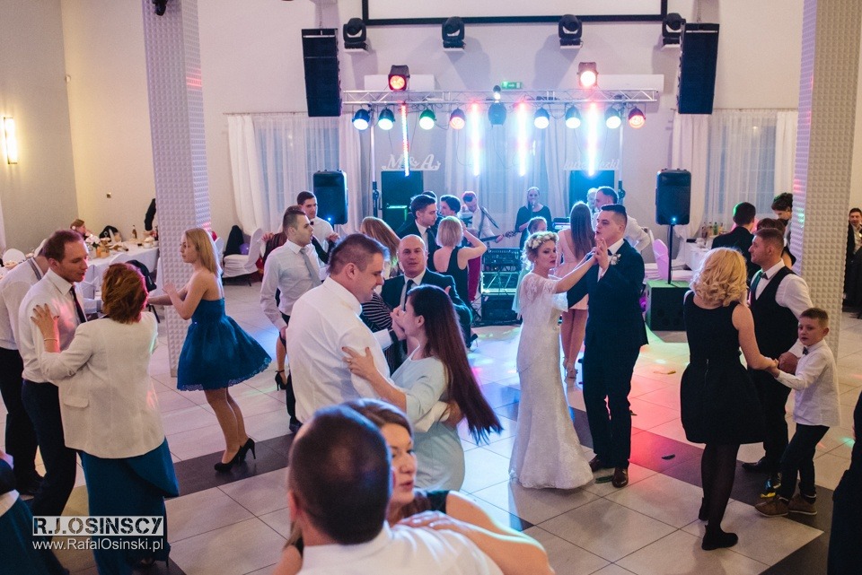 Goście weselni tańczą na parkiecie w sali balowej z oświetleniem scenicznym, widoczny zespół muzyczny w tle.